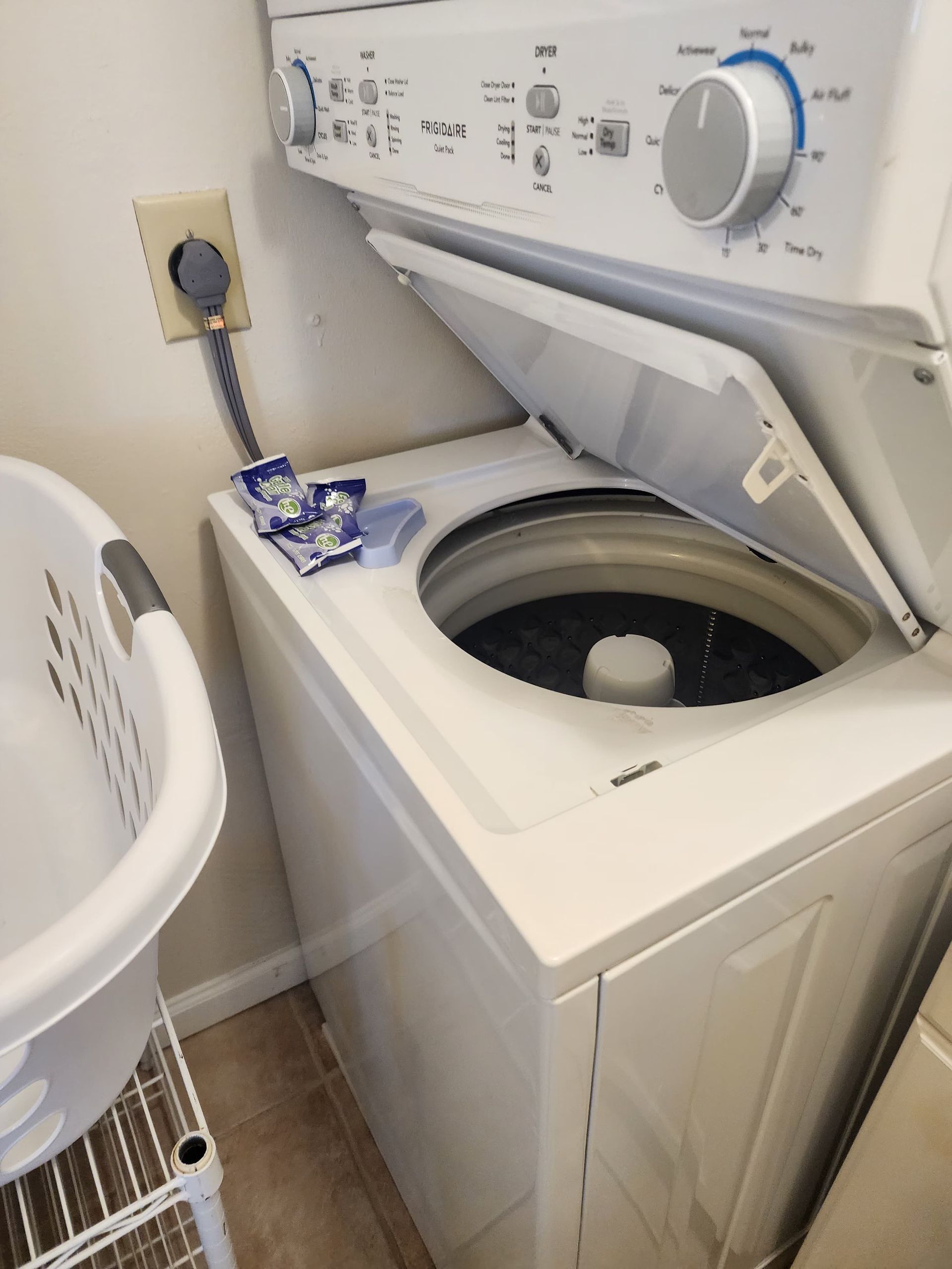 White stacked washer and dryer with open top; laundry basket and detergent pods nearby.