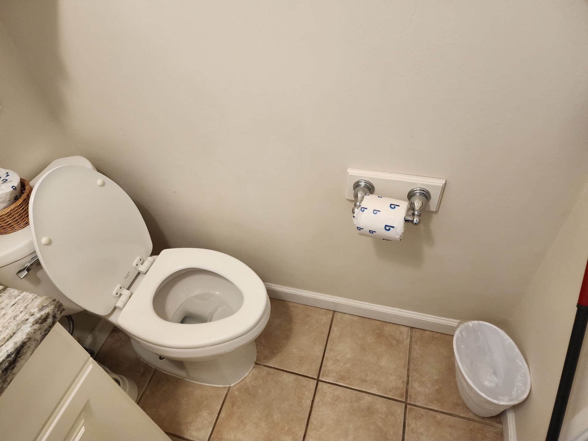 Toilet in a small bathroom with the seat up. There is a roll of toilet paper on the wall and a wastebasket on the floor.