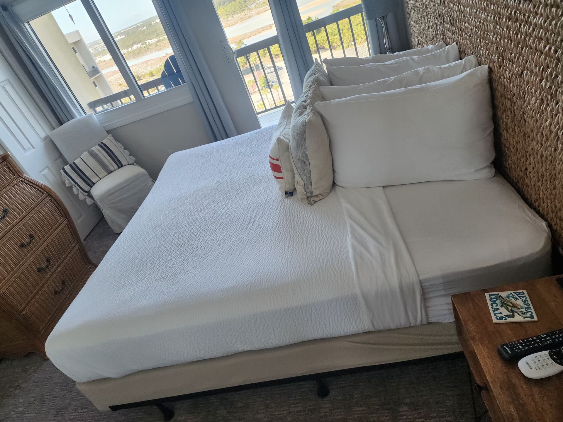 Bedroom with a made bed, white linens, pillows, wicker headboard, and wooden nightstand.