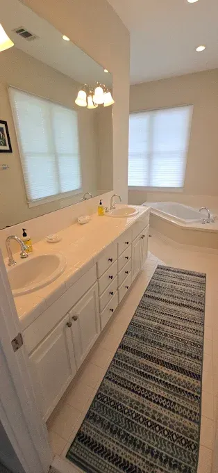 Bathroom with white cabinets, sinks, rug, and tub. Windows with blinds are visible.
