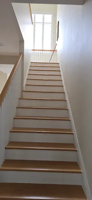 Wooden staircase leading upward, with a bright window at the top. White walls and railings.