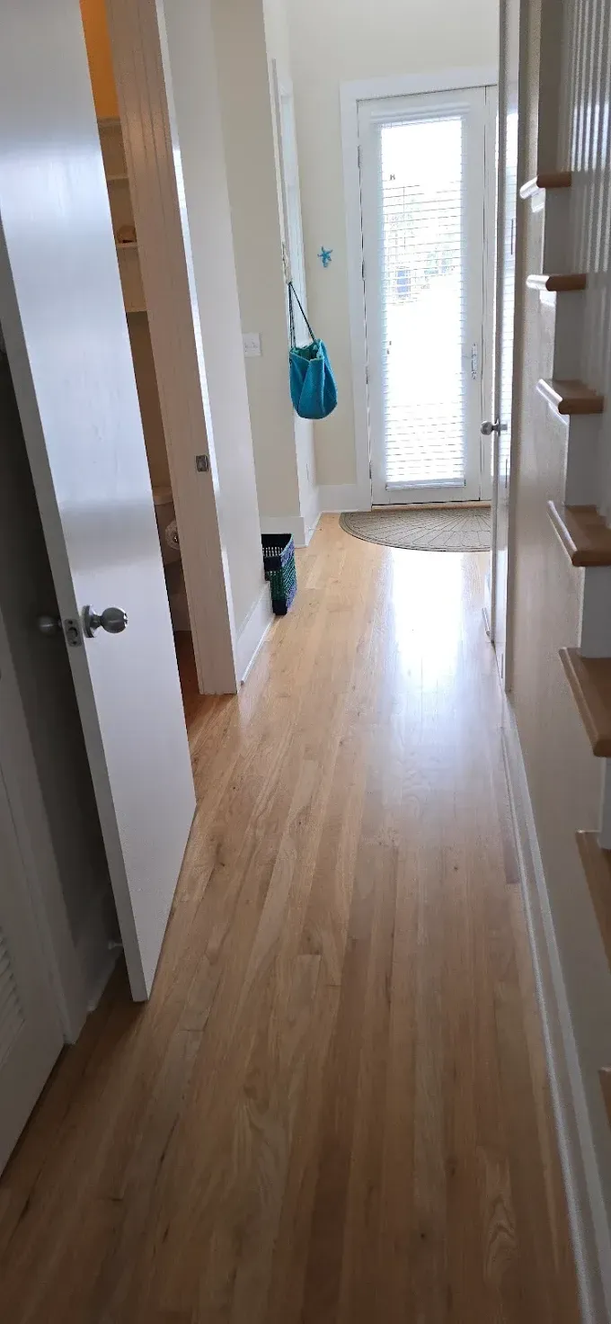 Hallway with wood floors, white walls, and a door at the end with natural light. A blue bag hangs on the wall.