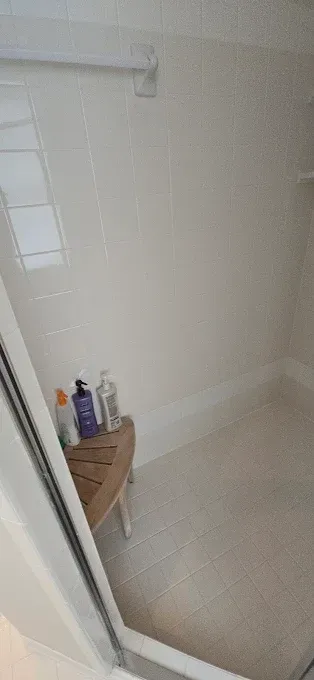 Inside view of a shower with a bench holding toiletries. White walls and a clear glass door are visible.