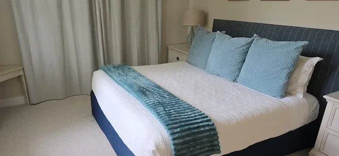 Bedroom with a white bed, blue pillows, headboard, and throw. Beige walls, curtains, and carpet.