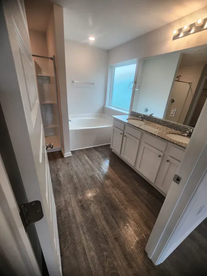 Bathroom with white cabinets, tub, and wood-look floor. A storage niche and large mirror are also present.