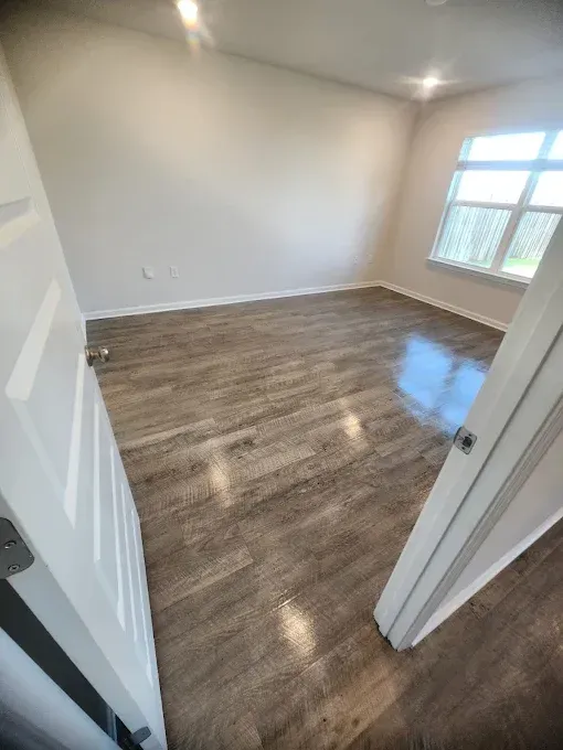 Empty room with gray wood-look flooring, white walls, and a window; the door is ajar.
