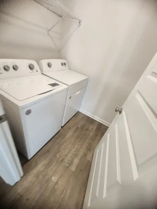 Laundry room with white washer, dryer, shelf, and door with wood-look flooring.