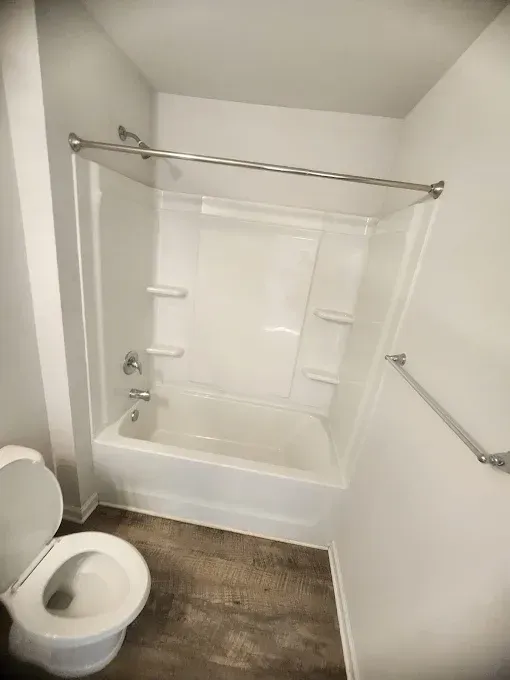 A small bathroom with a white tub/shower combo, toilet, and a towel rack. Gray walls and wood-look flooring.
