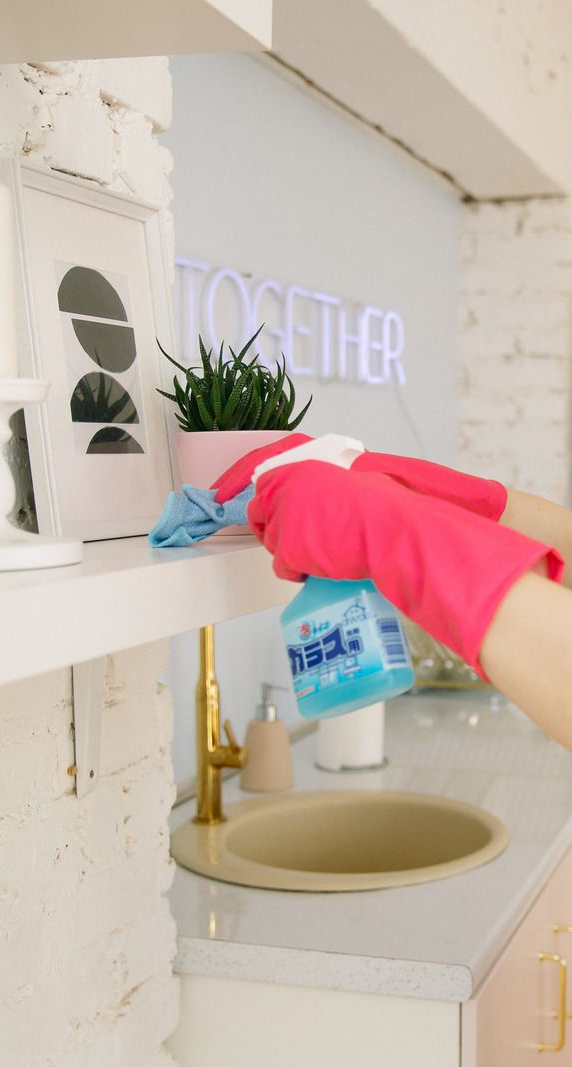 A person wearing pink gloves sprays cleaner and wipes a shelf in a kitchen with a pink neon sign.
