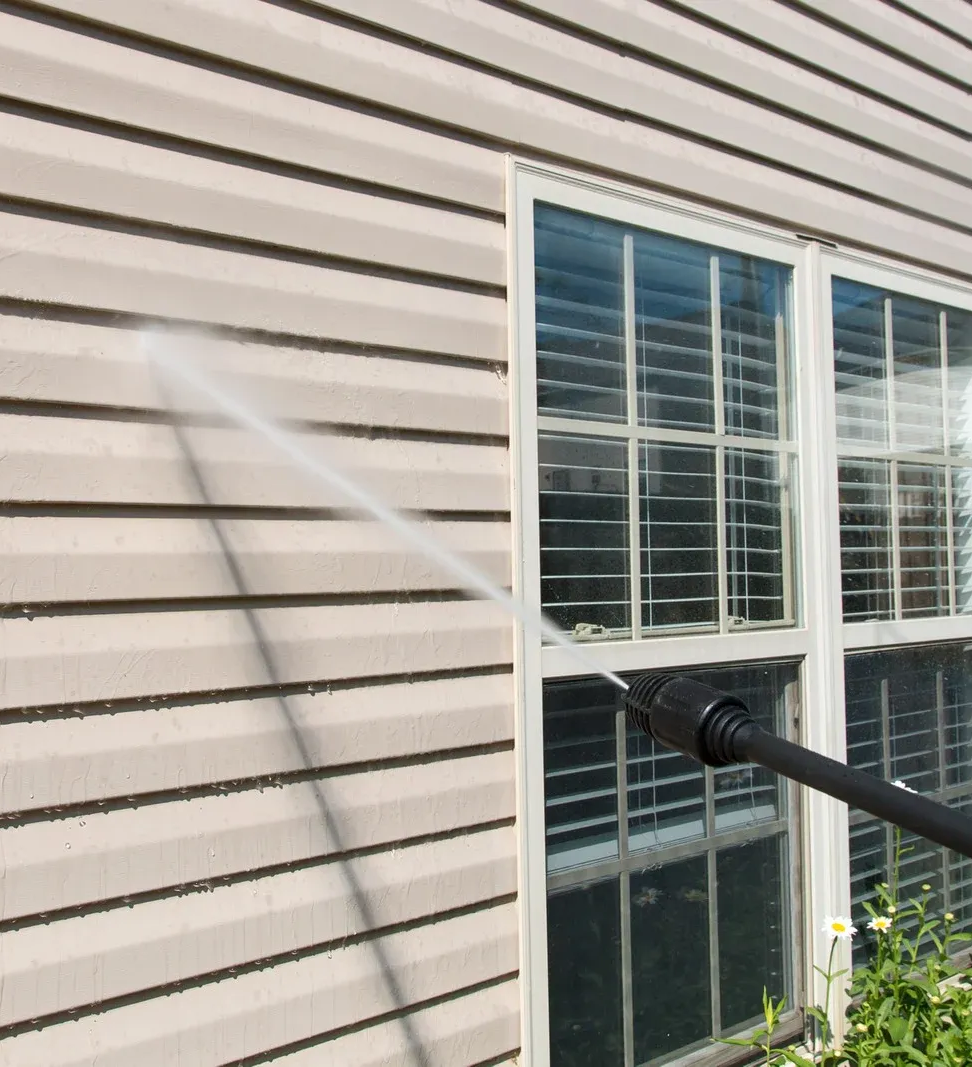 Pressure washing tan siding near a window. A stream of water sprays from a black nozzle.
