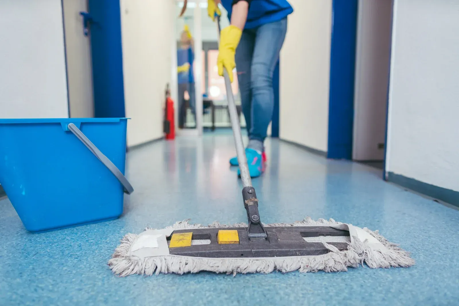 Person mopping a blue floor in a hallway, wearing yellow gloves. A blue bucket sits nearby.