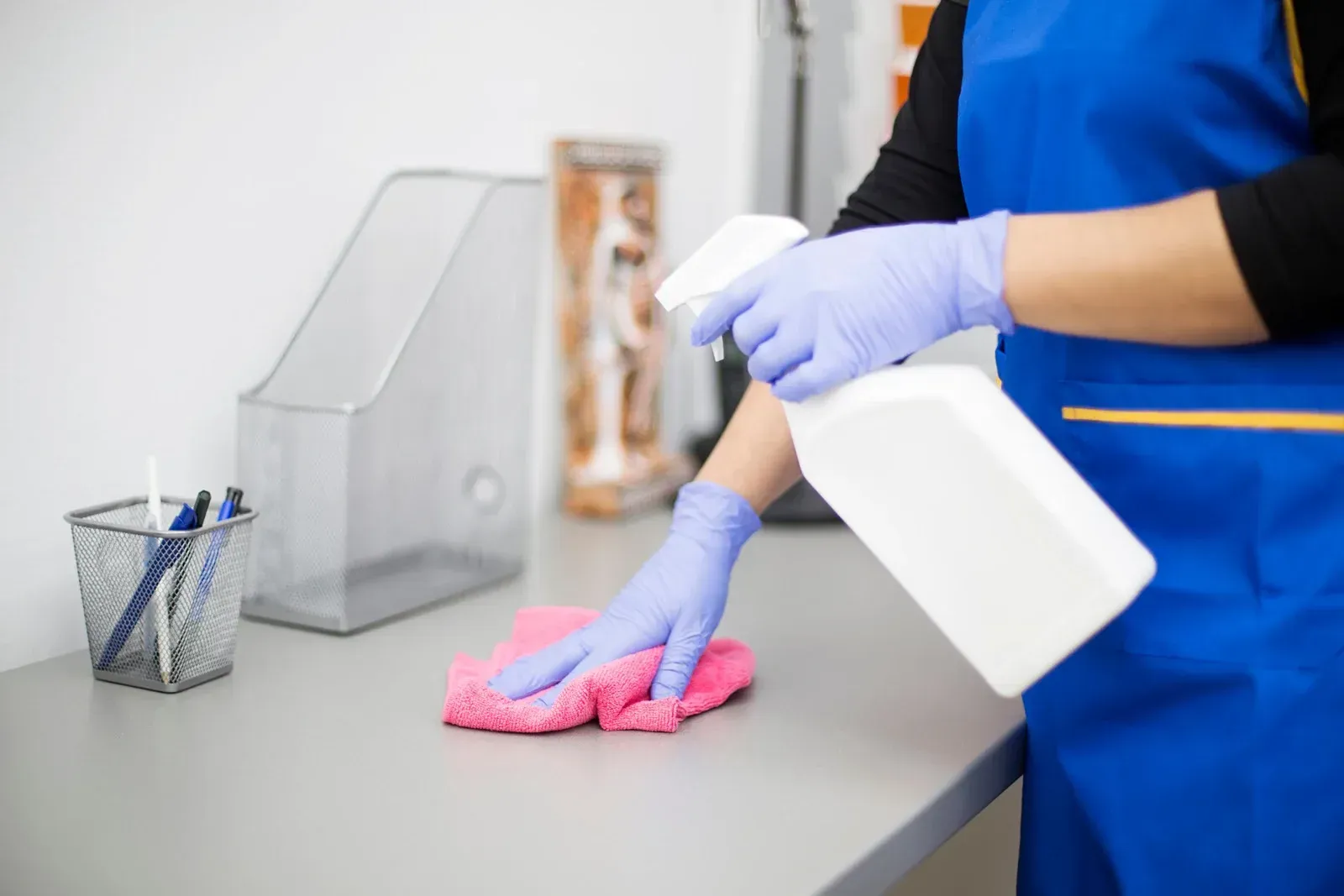 Person wearing gloves sprays and wipes down a gray desk, blue apron, pink cloth.