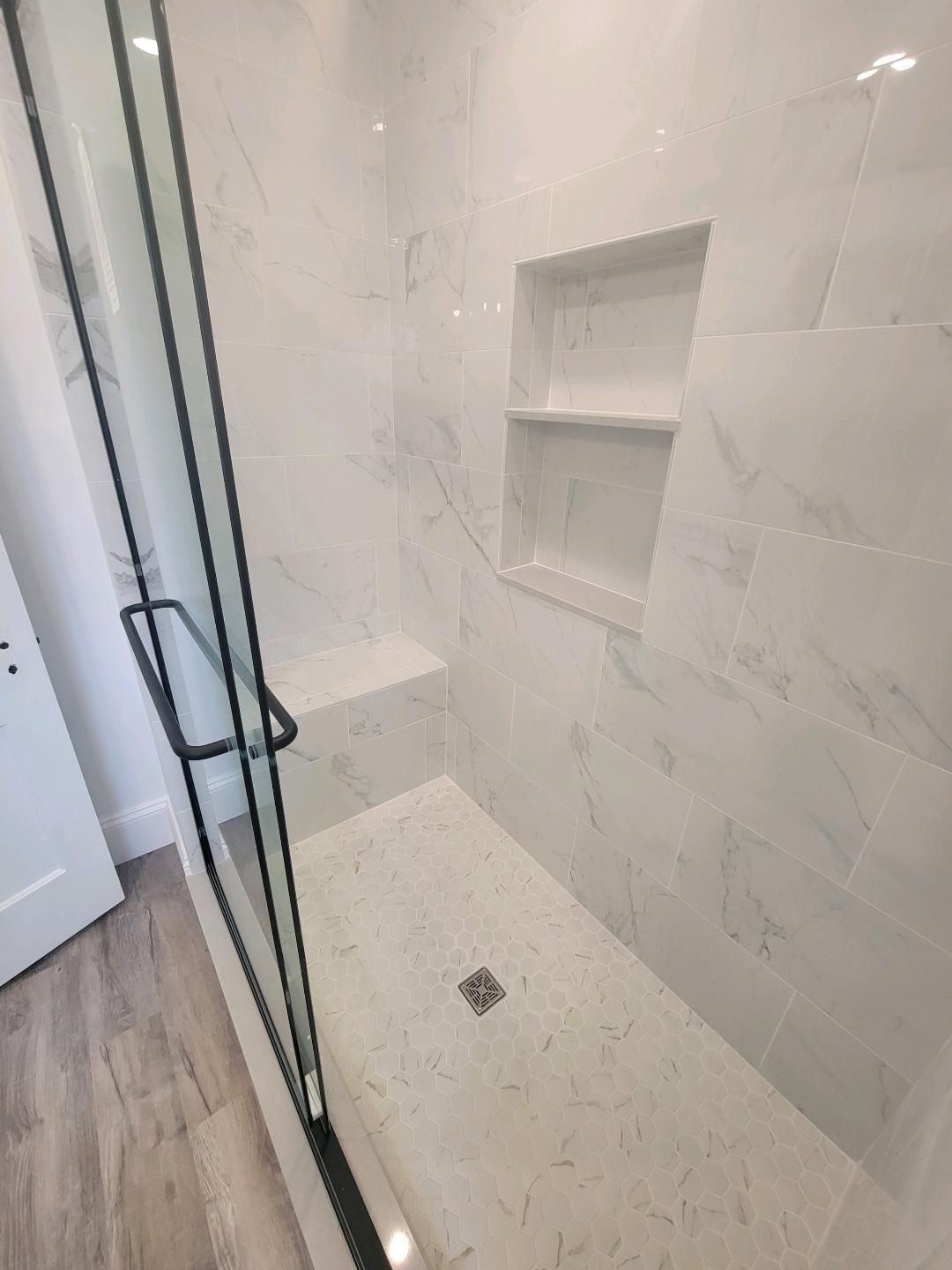 Marble-tiled shower with glass door, built-in bench and shelves. Black hardware, white walls, light wood floor.