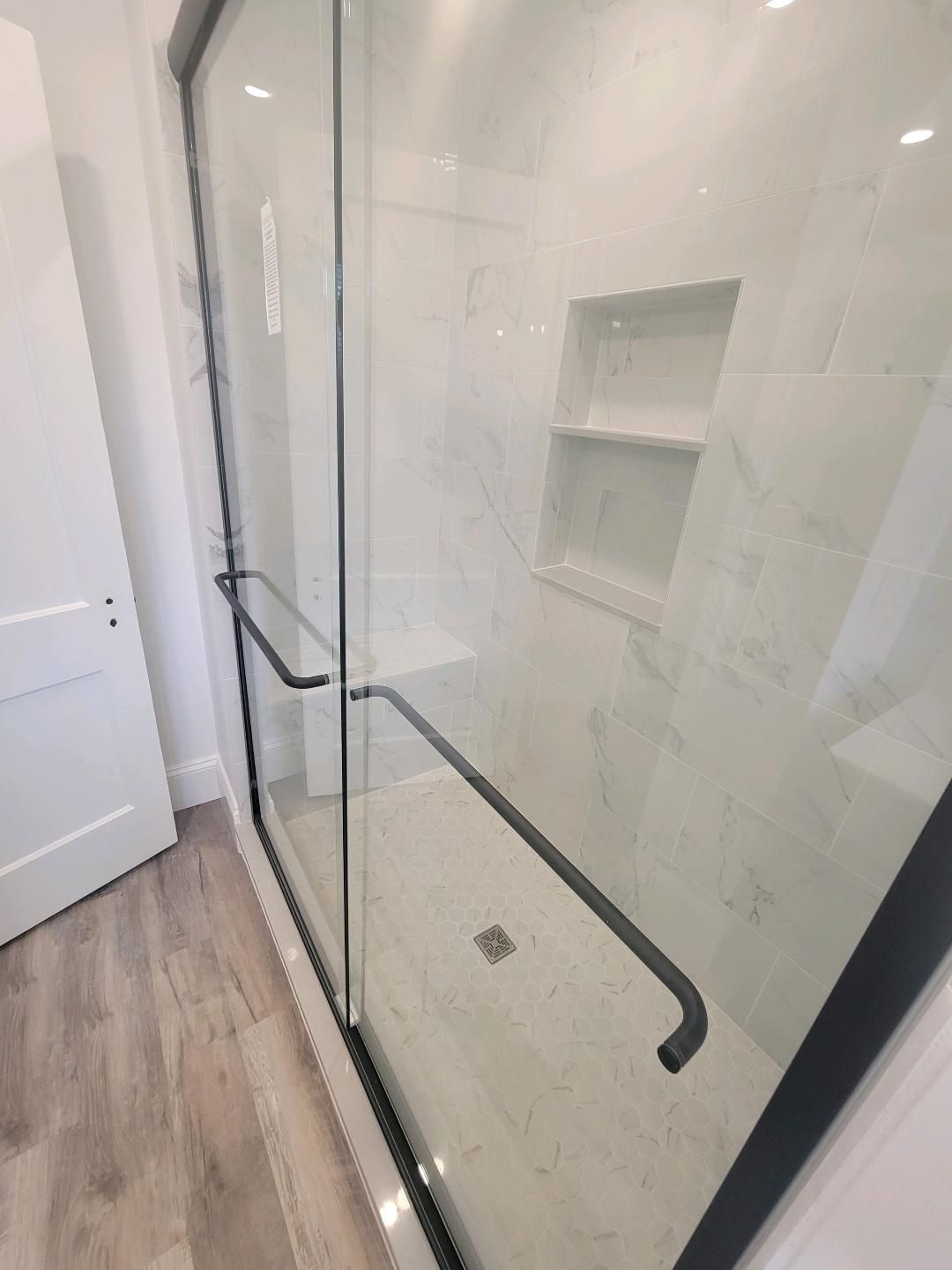 Glass shower with black handles, white tiled walls, and a recessed shelf.