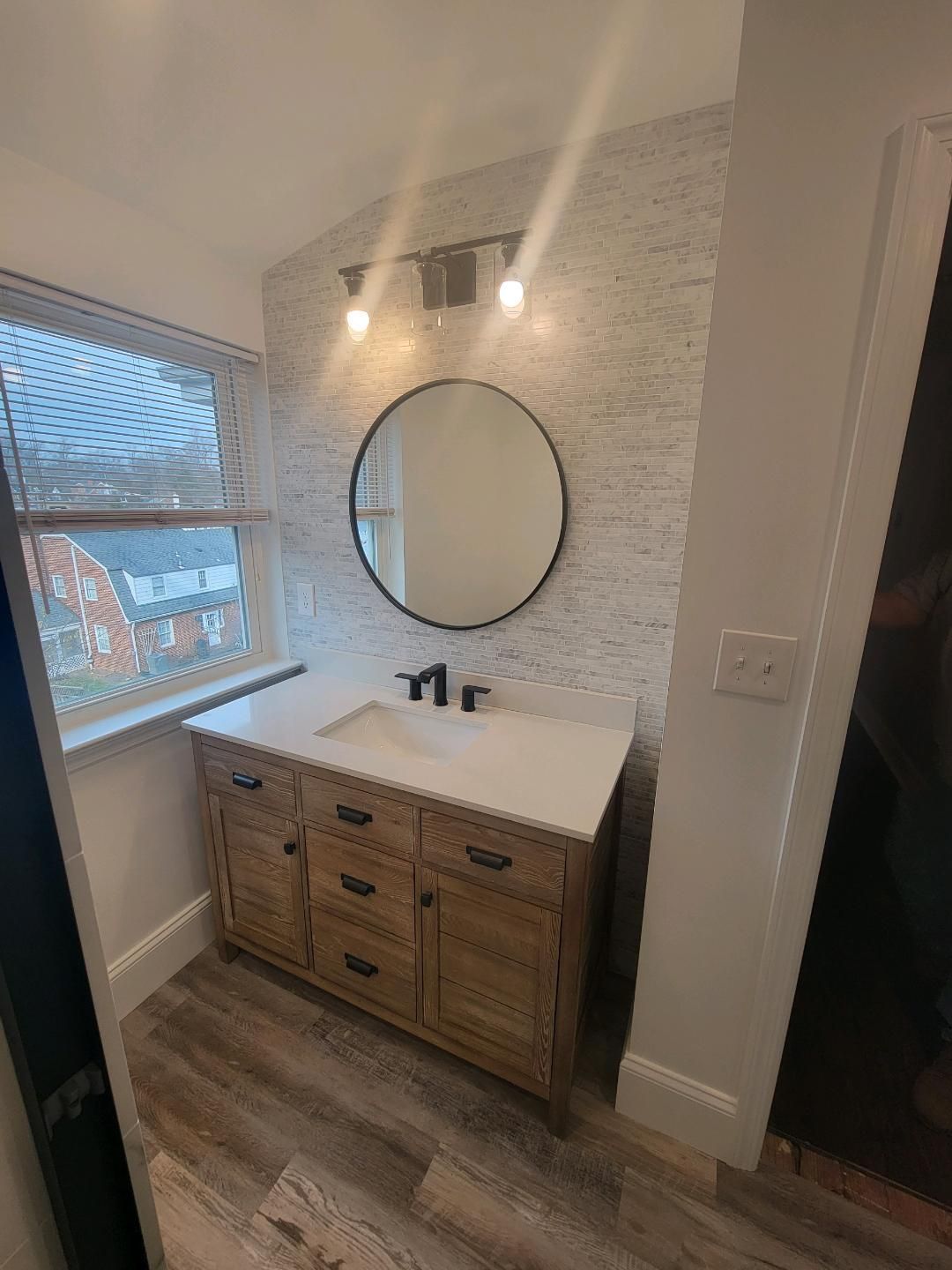 Bathroom with a wooden vanity, round mirror, white countertop, and stone-look wall behind the sink.
