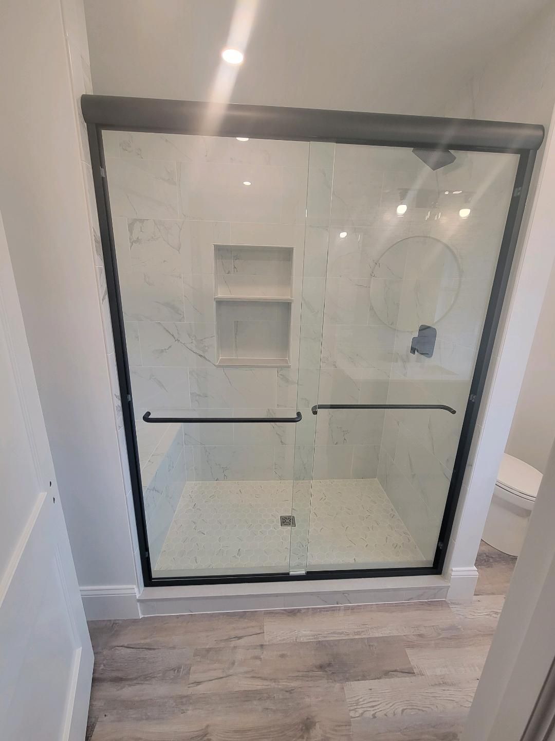 Black-framed glass shower with sliding doors, marble tile walls, and a built-in shelf in a bathroom with light wood flooring.