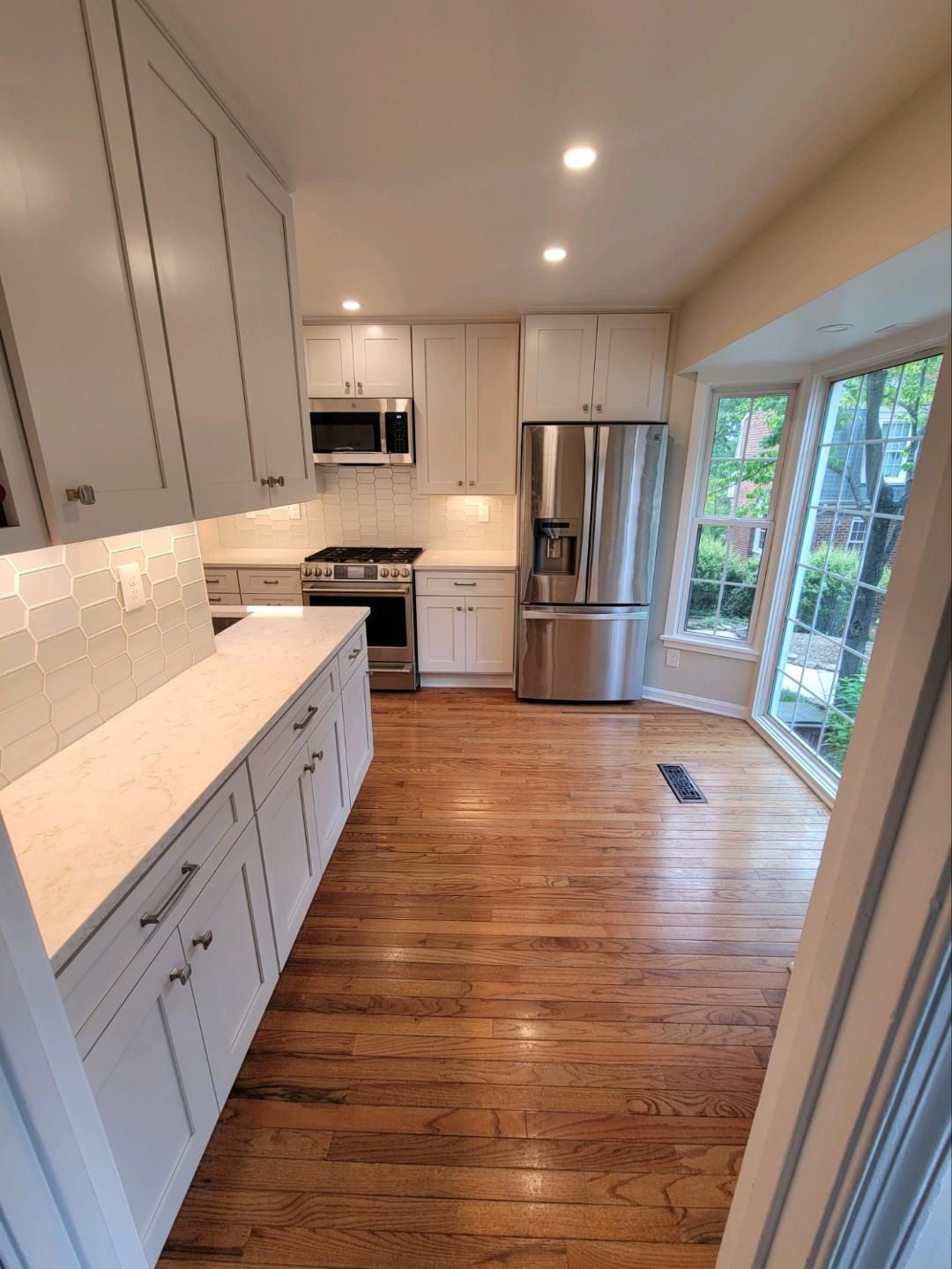 White kitchen with stainless steel appliances, light-colored countertops, and wood flooring. A large window brings in natural light.