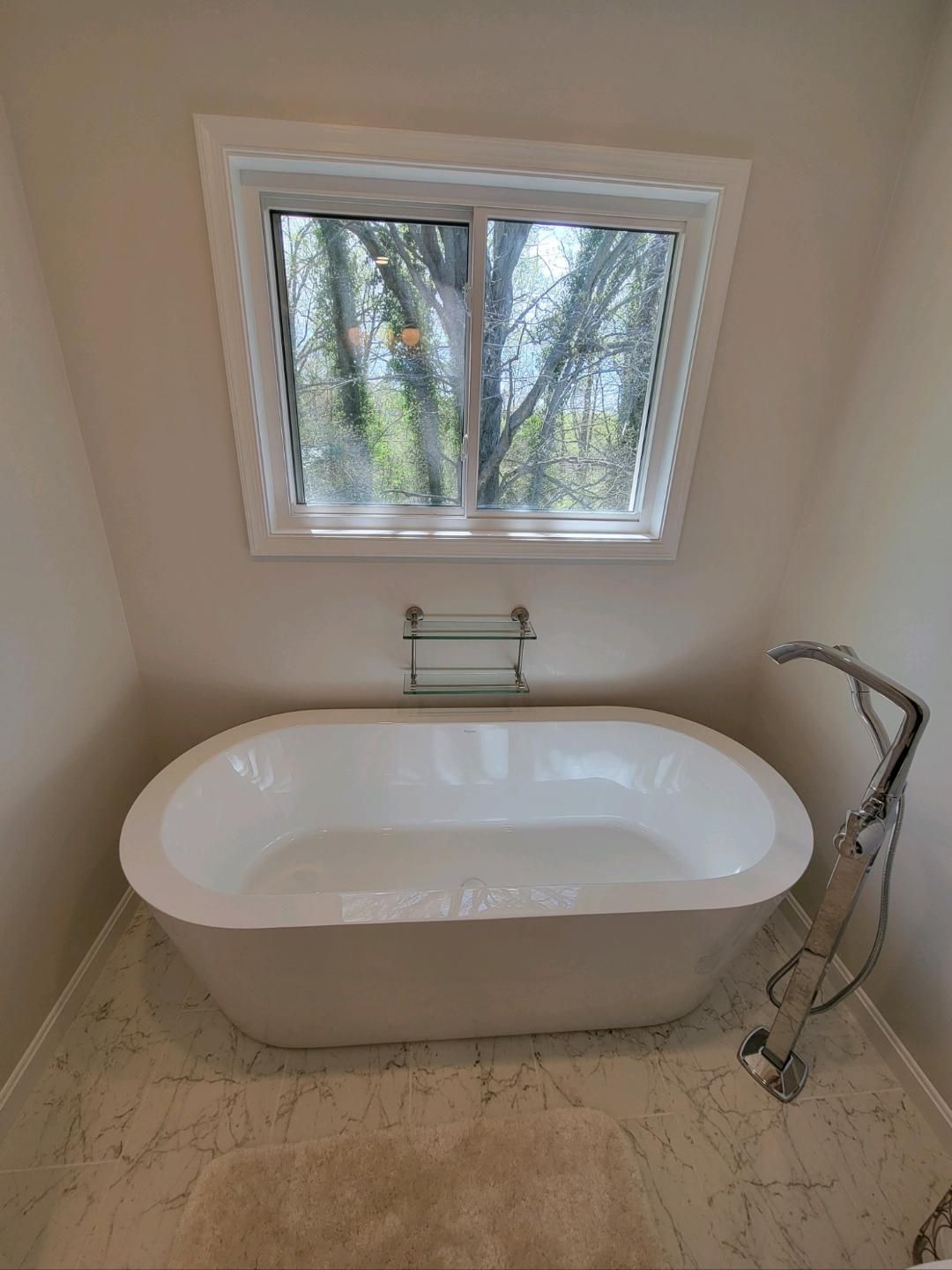 Freestanding white bathtub in bathroom with window and faucet; view of trees outside.