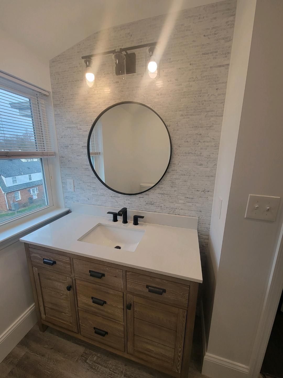 Bathroom with wood vanity, white countertop, round mirror, and textured accent wall.