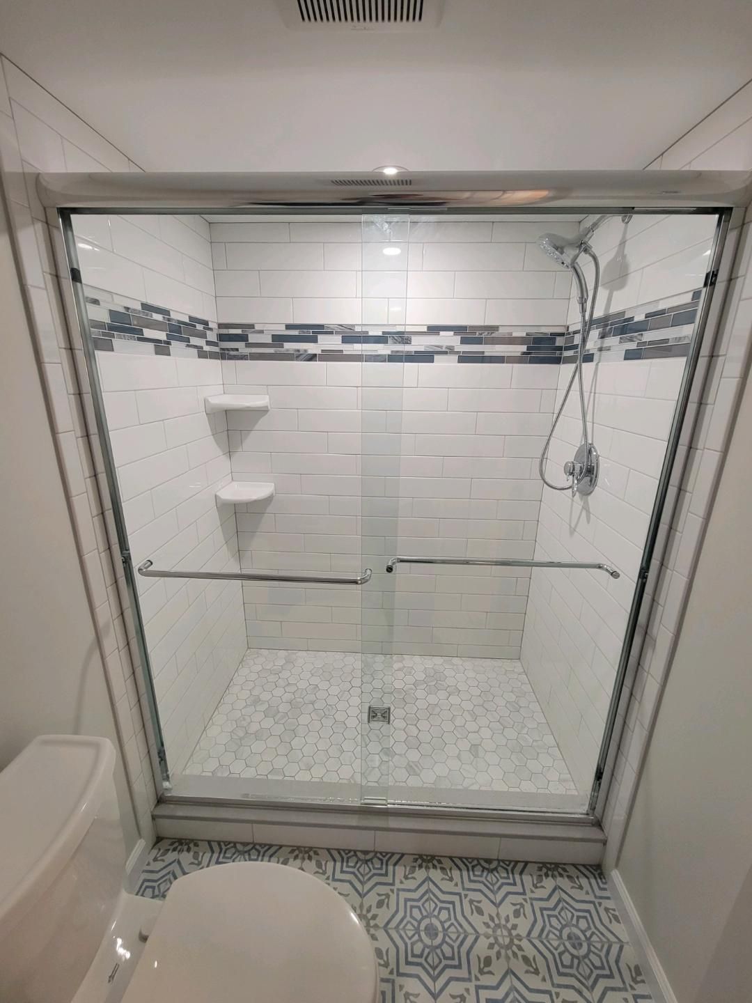 Shower with glass doors, white tile, decorative tile border, and a mosaic floor.