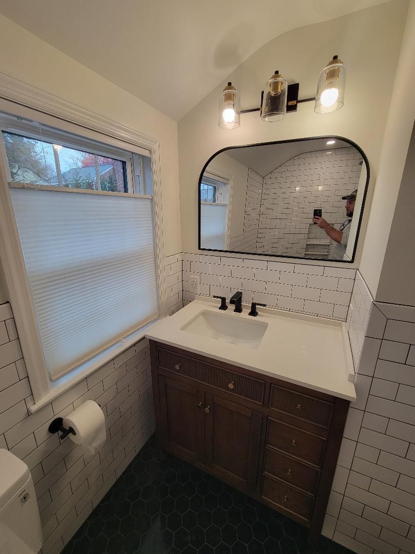 Bathroom with a dark wood vanity, white countertop, arched mirror, and textured white tiled walls.