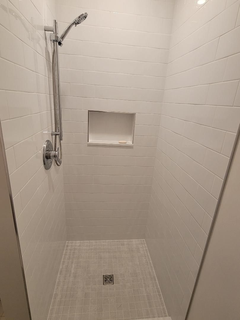 White tiled shower with a built-in shelf, showerhead, and floor drain.
