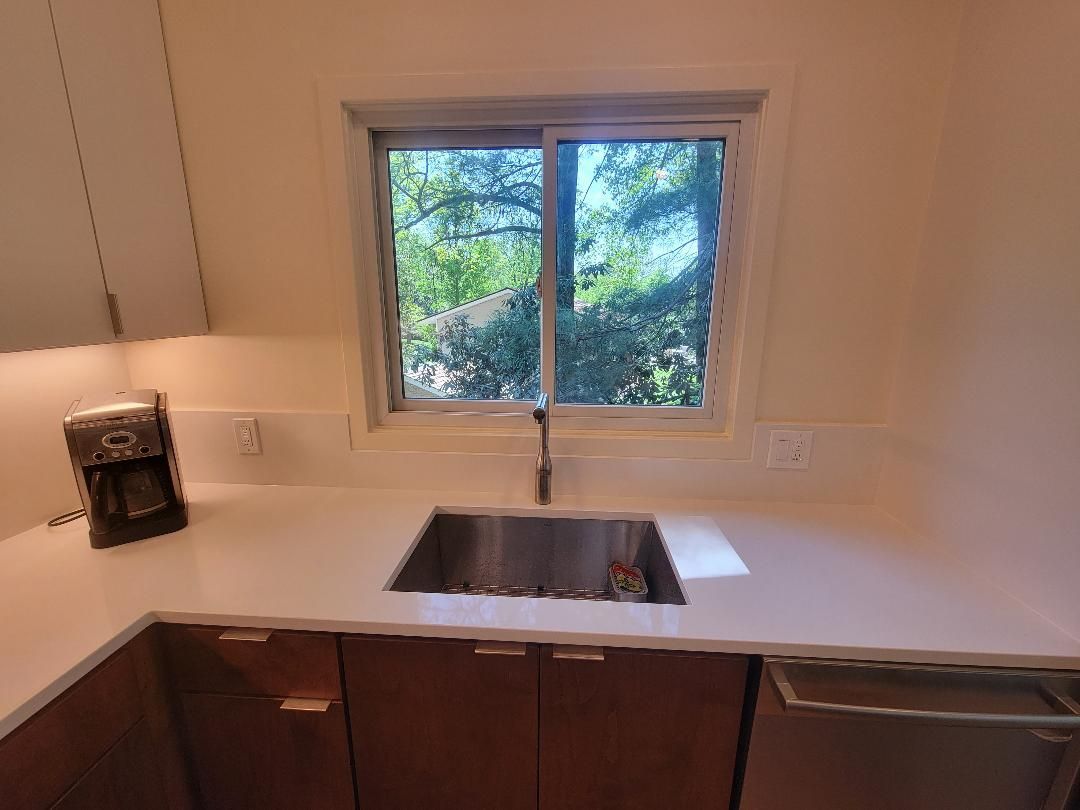 Kitchen with stainless steel sink, window with trees outside, white countertops, and brown cabinets.