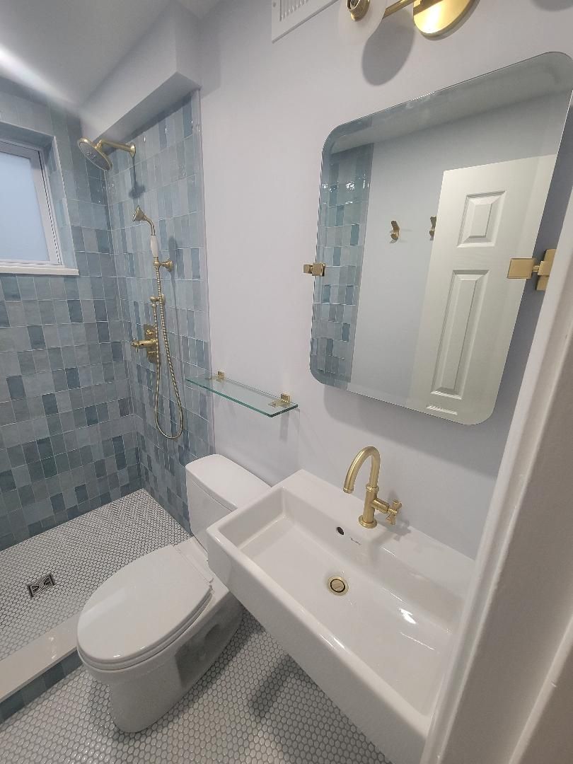 Small bathroom with blue tile shower, gold fixtures, white sink and toilet.
