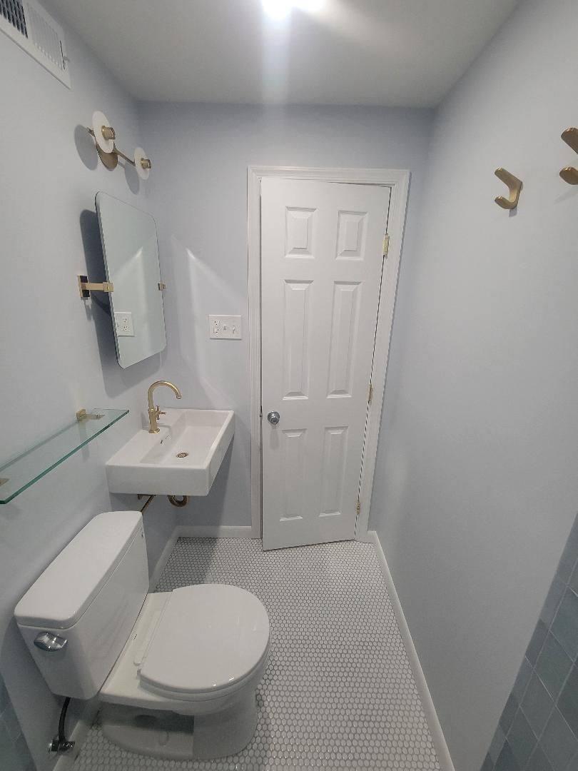 Small, light-blue bathroom with a toilet, sink, mirror, and closed door. Brass fixtures and white and gray patterned tile.
