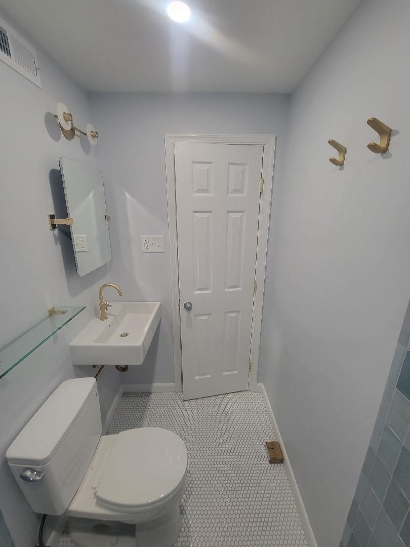 Small, light-blue bathroom with a white toilet, sink, and door; gold fixtures; a patterned floor; and wall hooks.