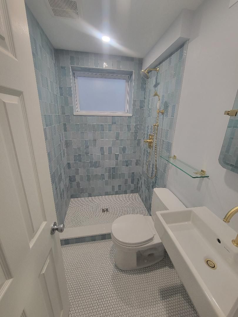 Small bathroom with blue tiled shower, white toilet and sink, gold fixtures.