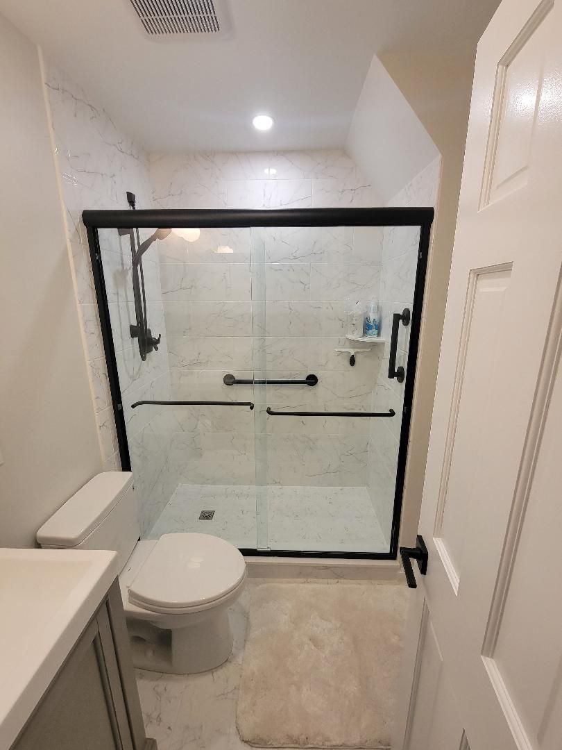 Bathroom with a black-framed shower, toilet, and gray vanity. White walls and a light beige floor.
