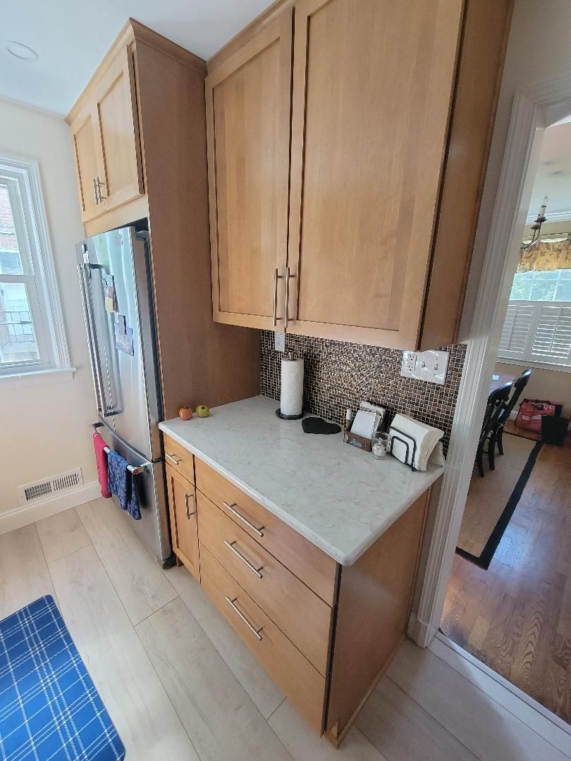 Kitchen with light wood cabinets, white countertop, and a patterned backsplash. Refrigerator on the left, doorway on the right.