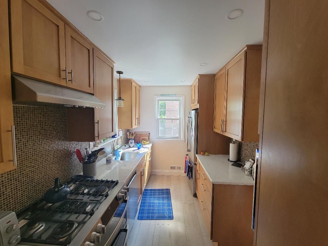 Narrow kitchen with light wood cabinets, white countertops, and a window at the end.