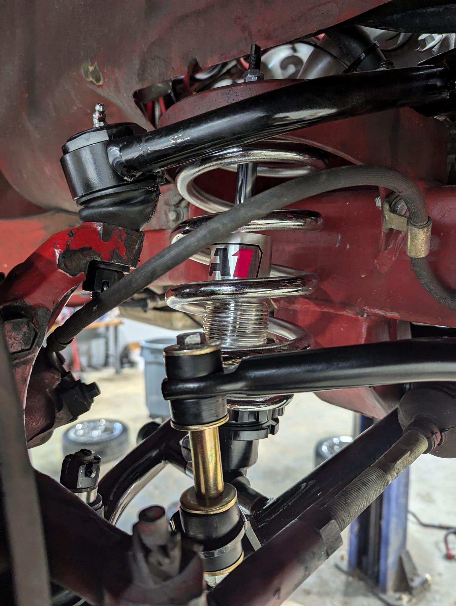 Car suspension assembly with black control arms, coil-over shock, and red bodywork.