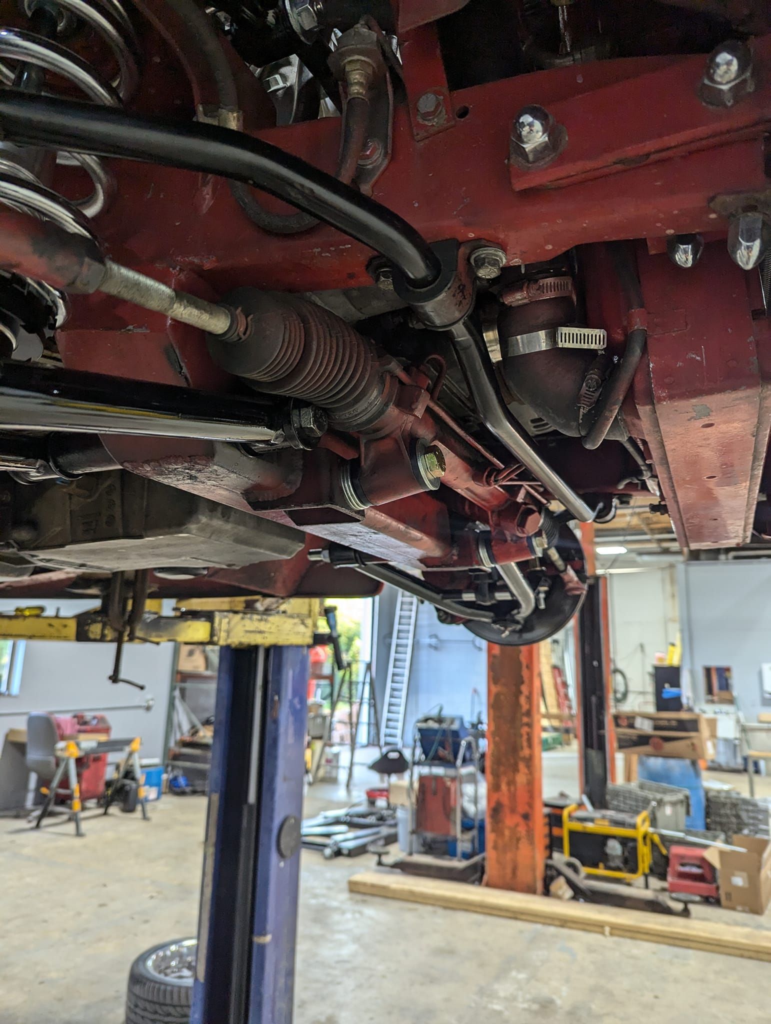 Undercarriage of a red car on a lift, showing steering components and suspension in a workshop.