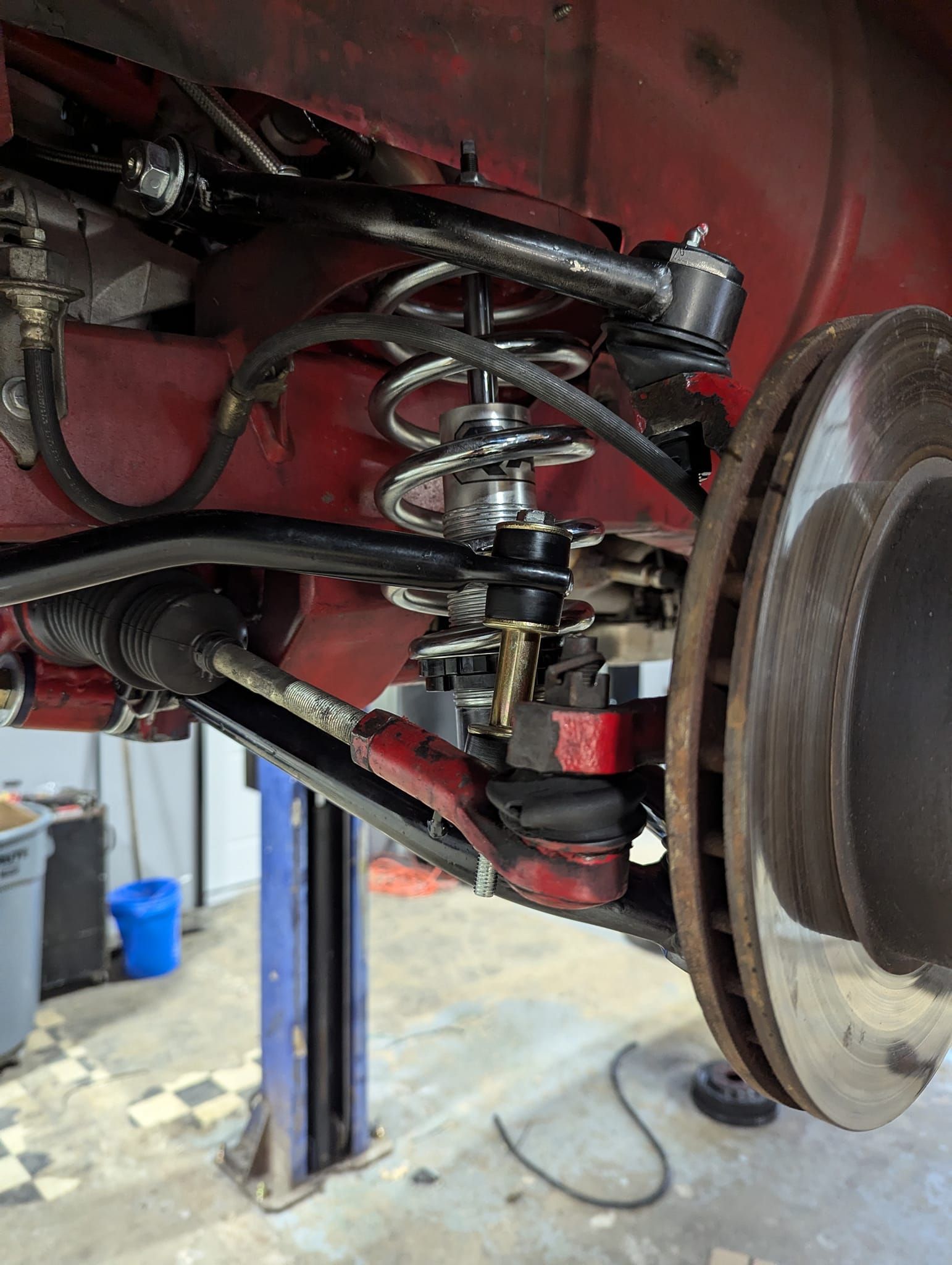 Red car's front suspension on a lift, showing coil spring, shock, upper/lower control arms, and brake rotor.