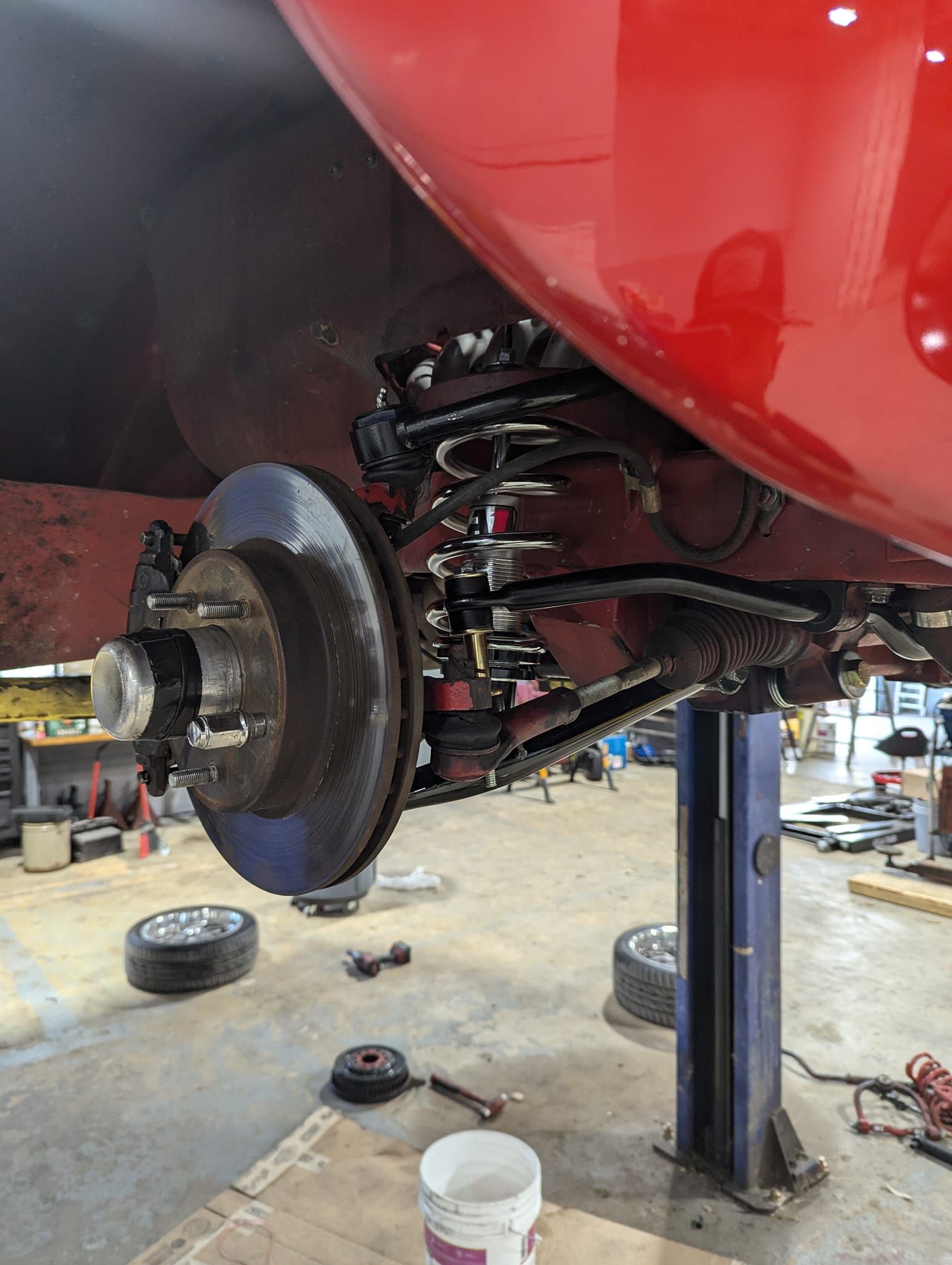 Red car's rear wheel and suspension elevated on a lift in a garage, showcasing brake disc and coil springs.