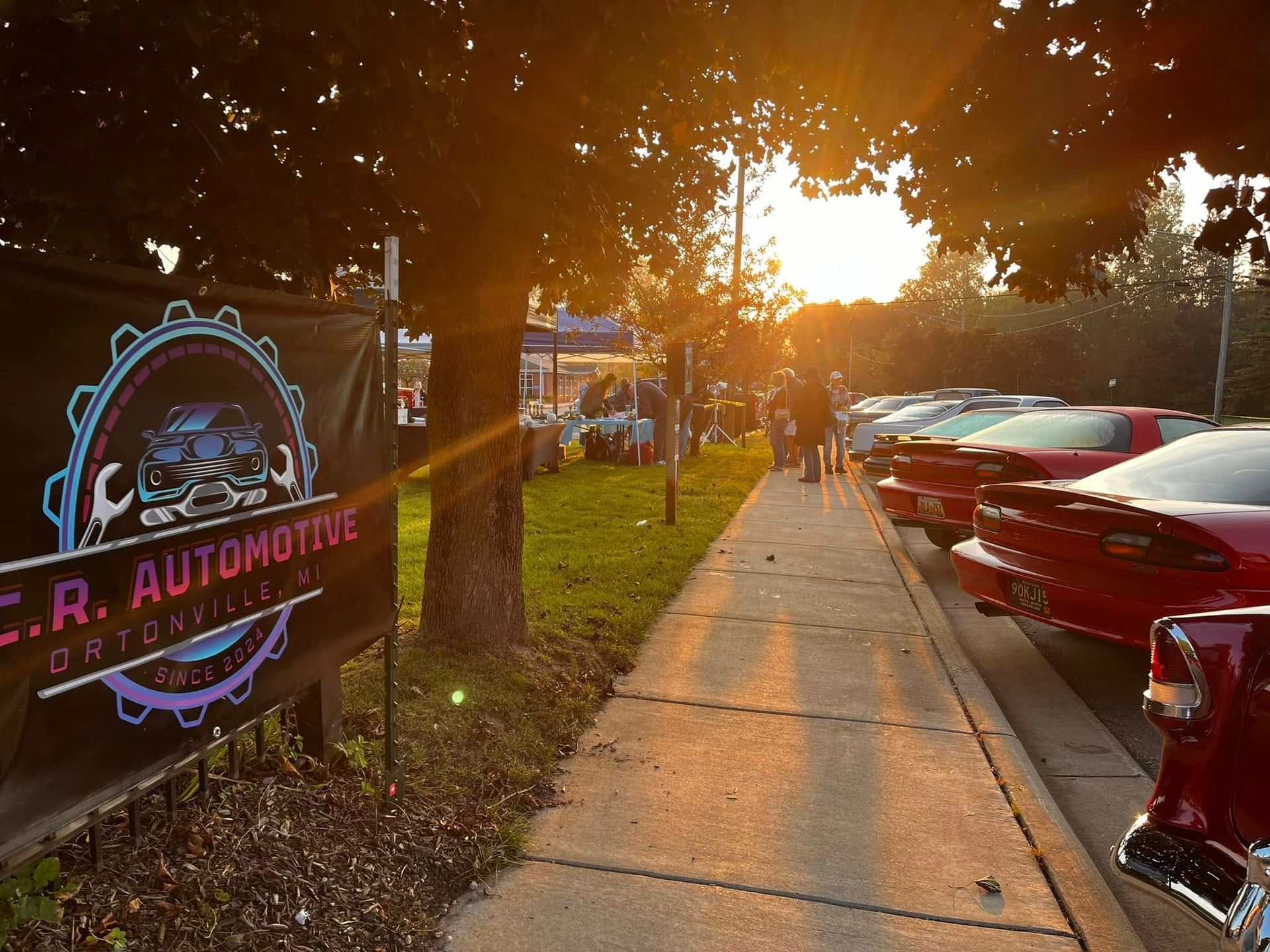Car show at sunset with banner for C.R. Automotive. Cars line the sidewalk.