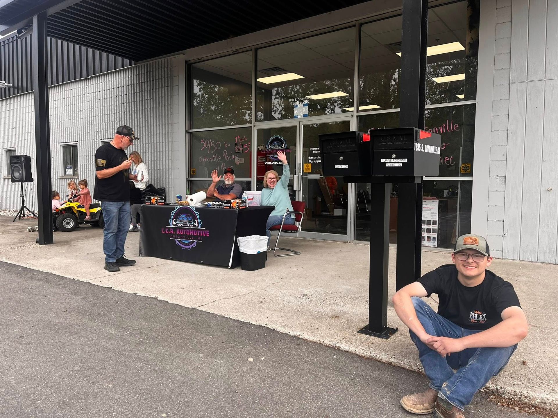 People outside a building with a table; one waves. Black mailbox and speaker visible.