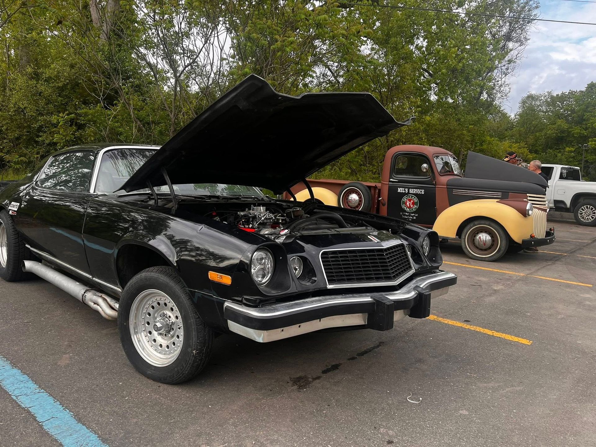 Black classic Camaro with hood up, parked next to a vintage red truck.