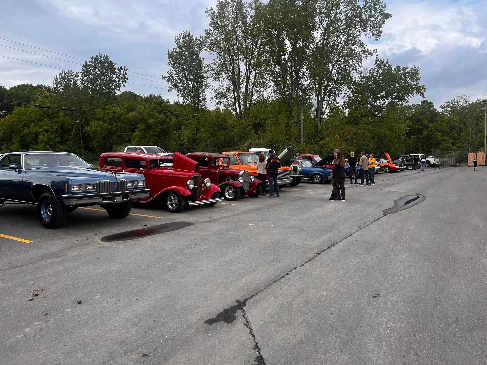 Classic cars parked in a lot, people standing near them. Trees and overcast sky in the background.