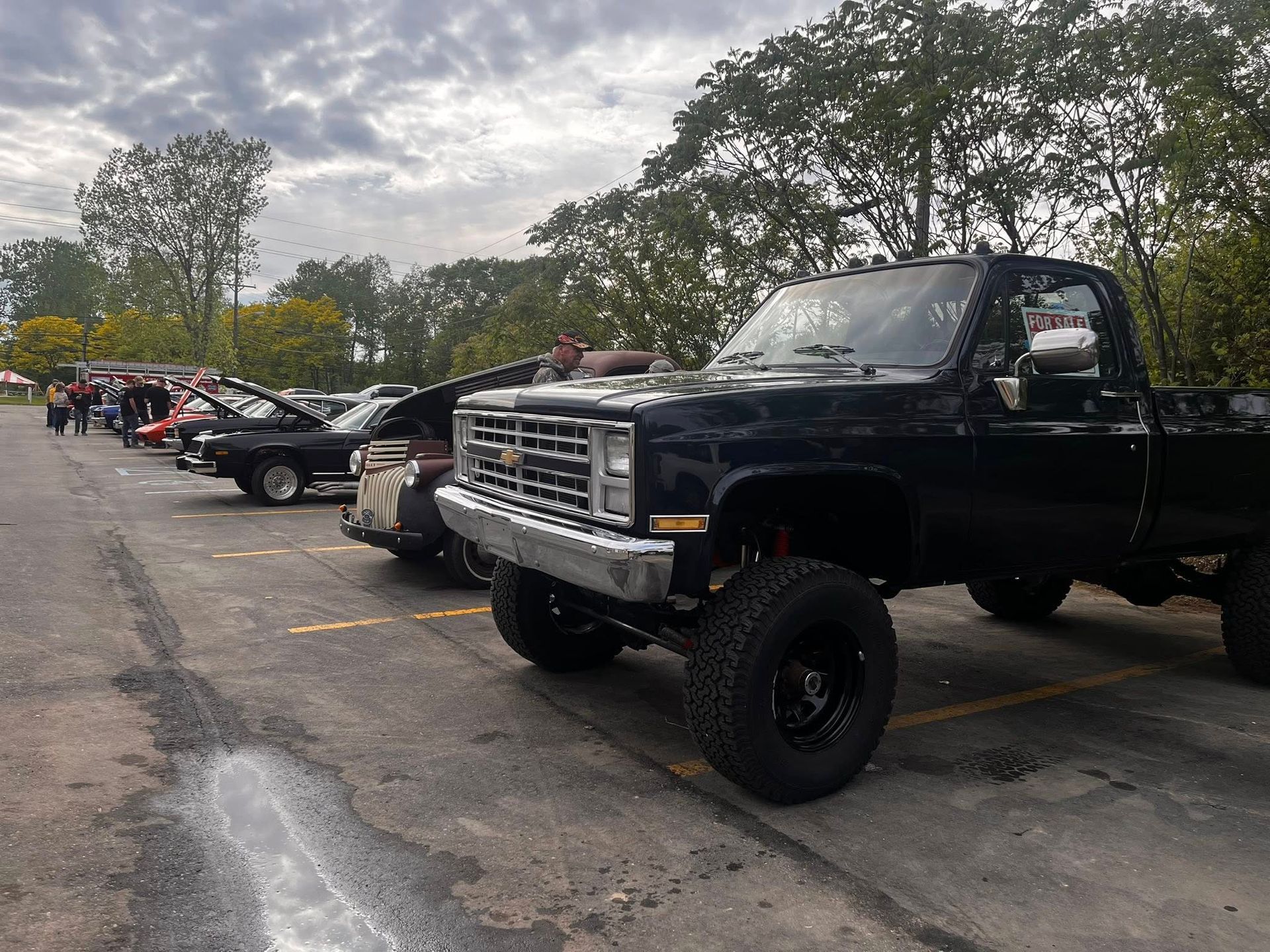 Black lifted pickup truck at a car show, parked in a lot. Other classic cars in the background. Cloudy day.