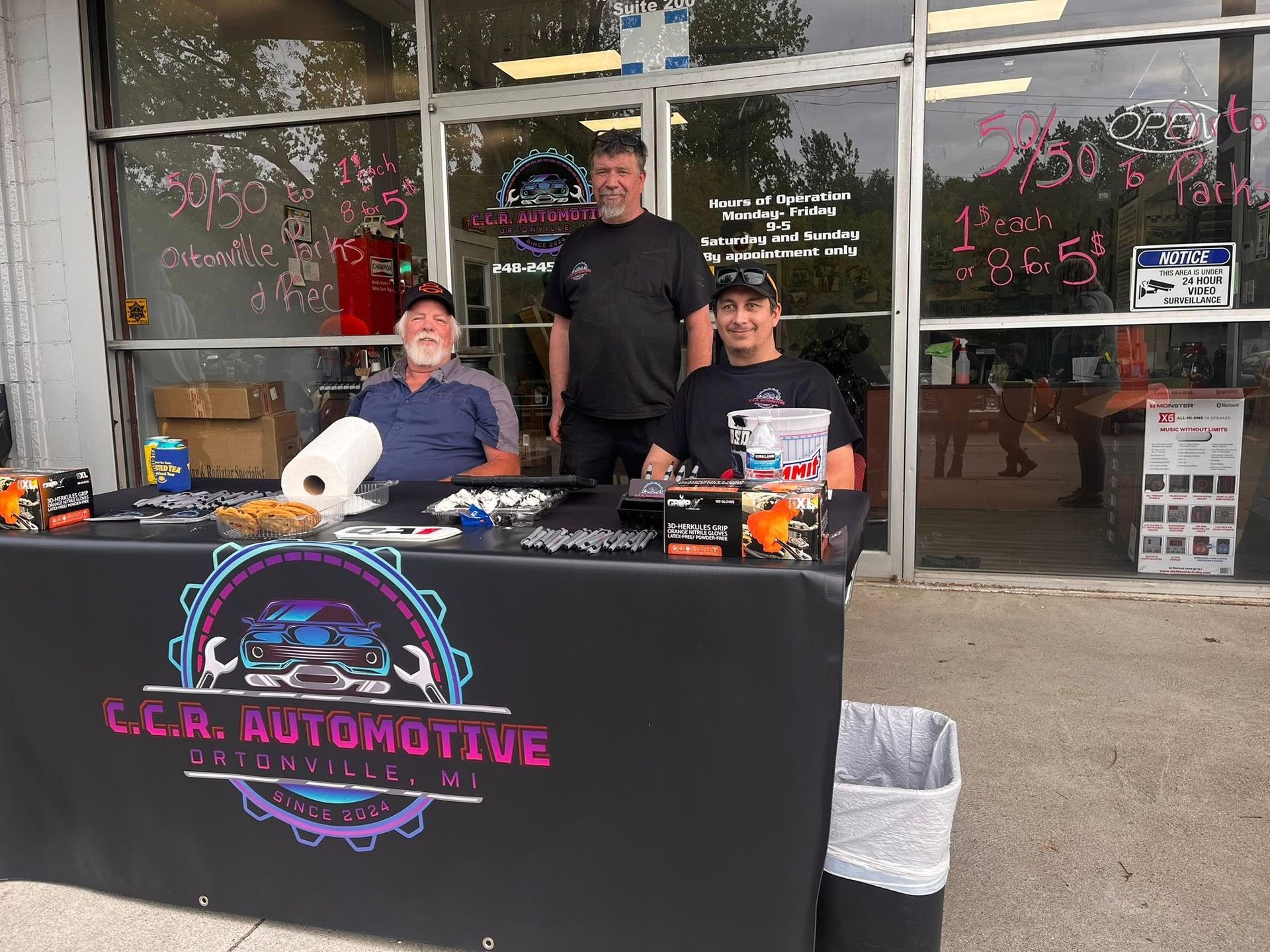 Three people stand behind a table promoting C.C.R. Automotive in front of a storefront.