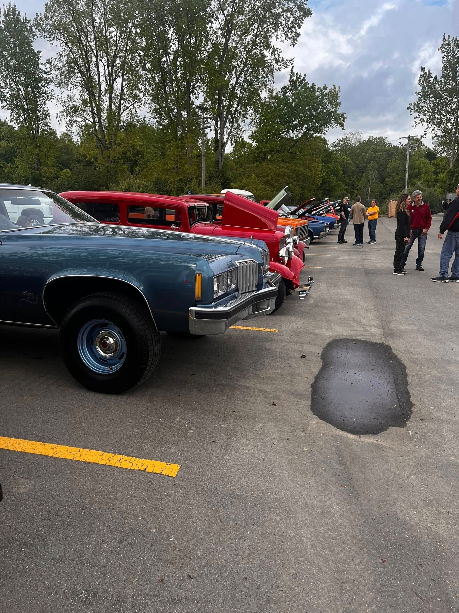 Classic cars parked in a lot, people milling around. Blue car in the foreground, red ones behind. Overcast sky.