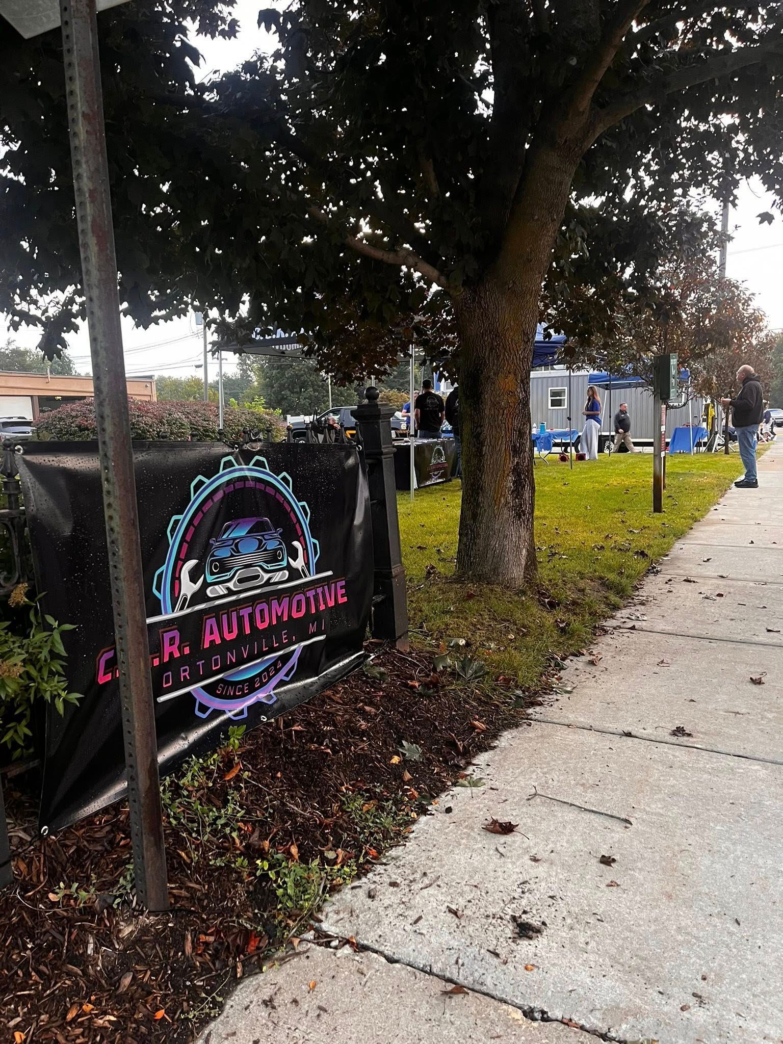 Sign for C.R. Autohaus, featuring a car image, set outdoors with a sidewalk, grass, and a tree.