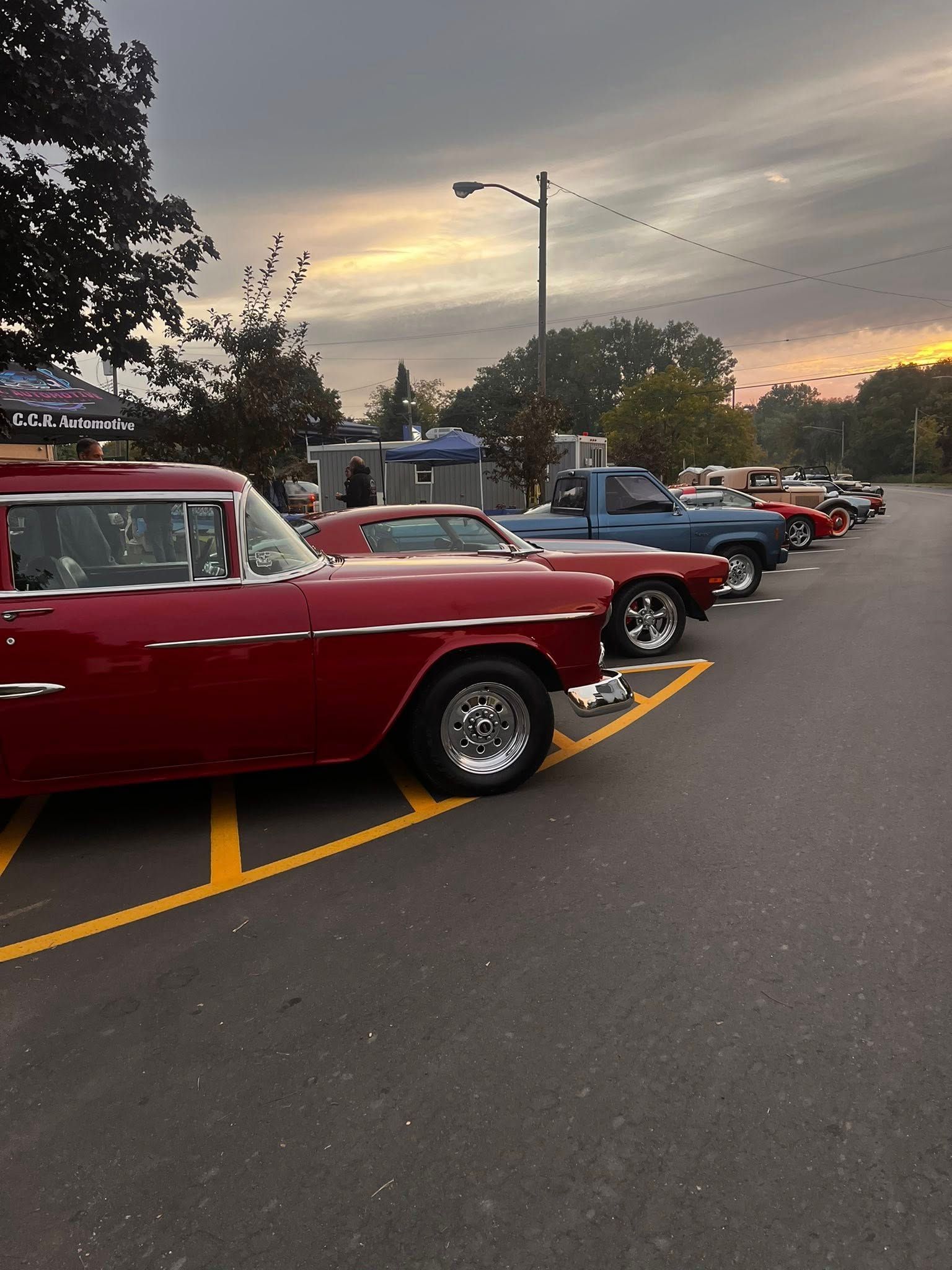 A row of classic cars in a parking lot at dusk. Red, blue, and brown vehicles are parked in a line.