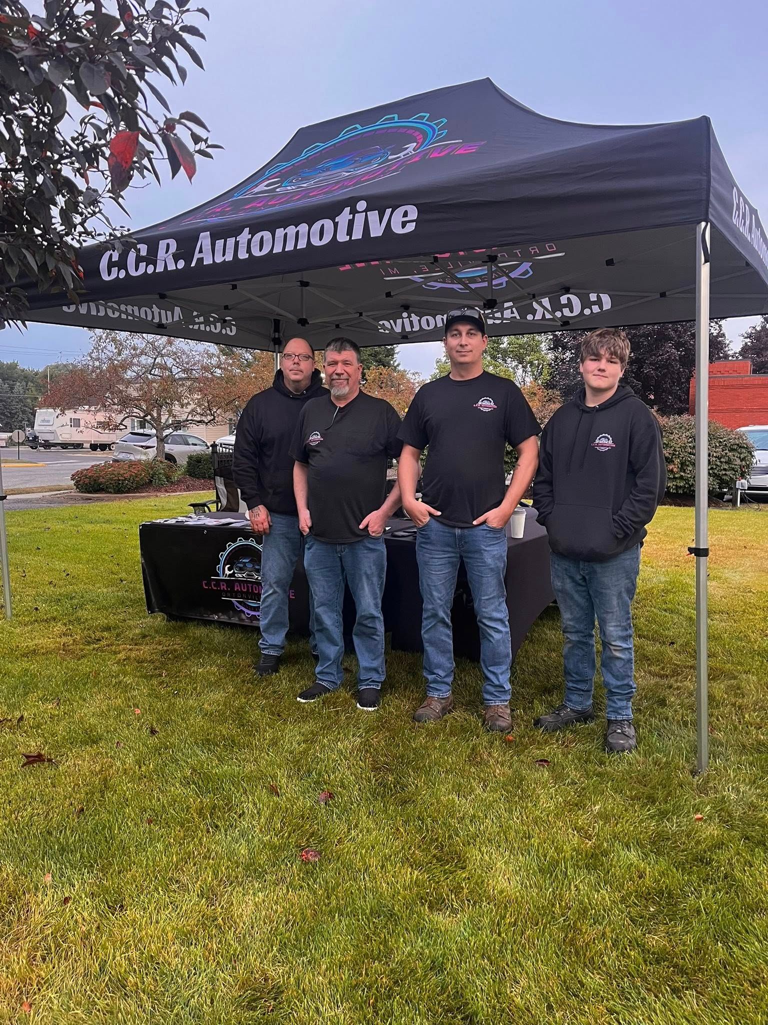 Four men standing under a tent with “C.C.R. Automotive” logo. They are on grass with vehicles in the background.