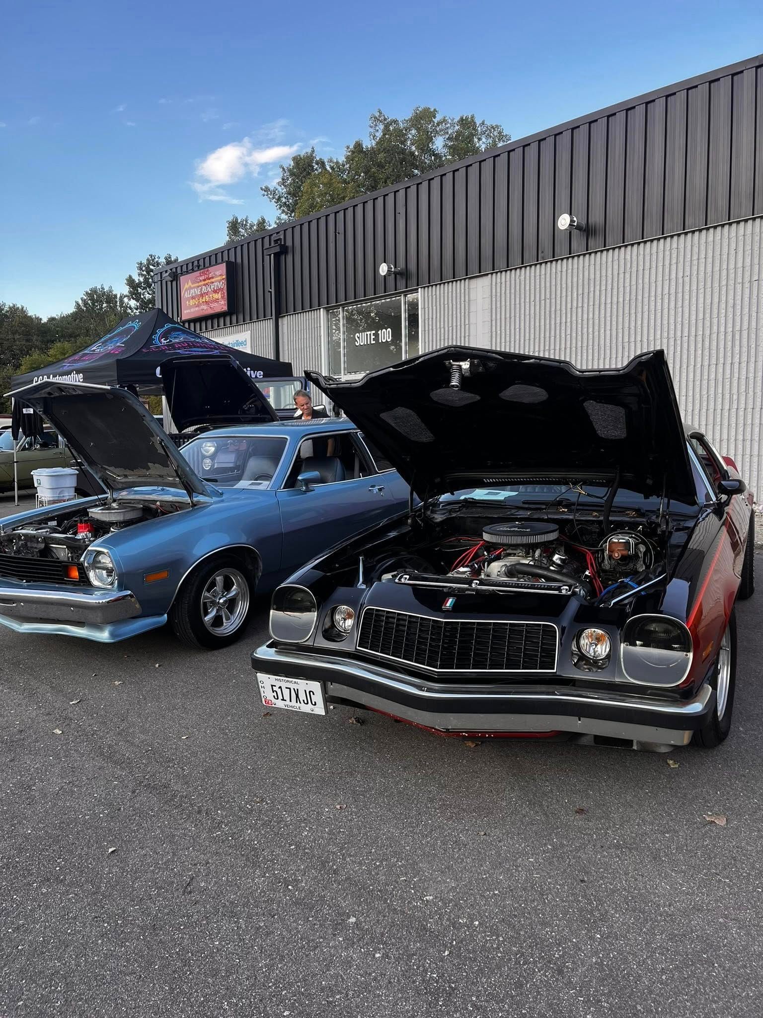 Two classic cars with open hoods parked outside a building on a sunny day.