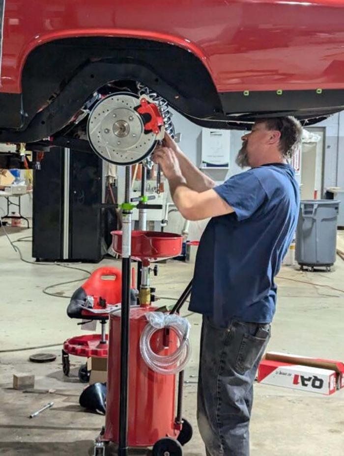 Man working on the brakes of a red car in a garage, using tools. Oil drain on wheels is nearby.